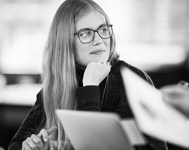 A woman wearing glasses sits at a table, focused on her laptop, with a calm expression in a well-lit room.
