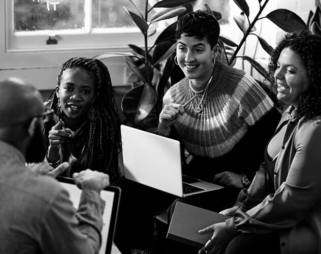 A diverse group of individuals sitting around a table with laptops, engaged in a meeting or collaboration session.