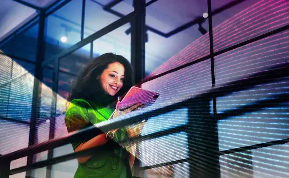 A woman stands in front of a building, holding a tablet and looking engaged with her surroundings.