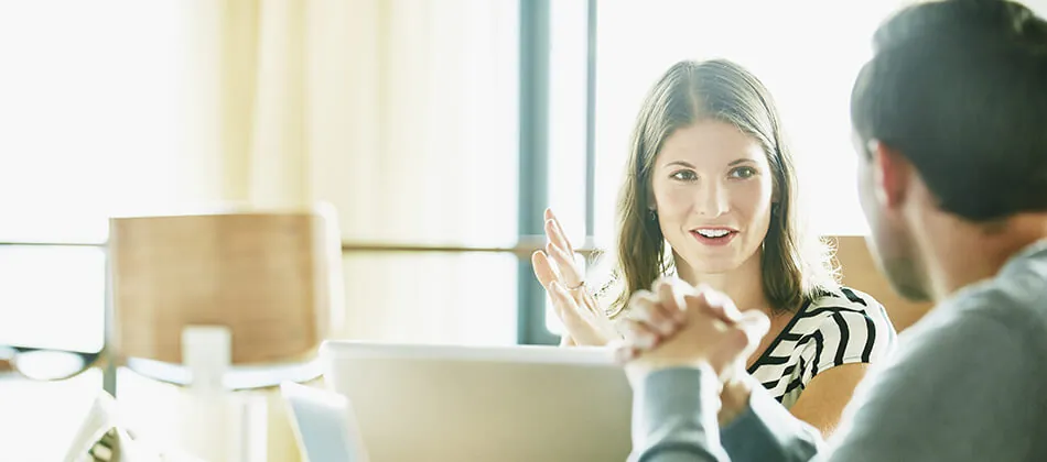 A woman and a man seated at a table, engaged with a laptop in front of them, focused on their work or discussion.