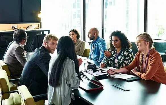 A diverse group of professionals engaged in discussion around a large conference table in a modern meeting room.