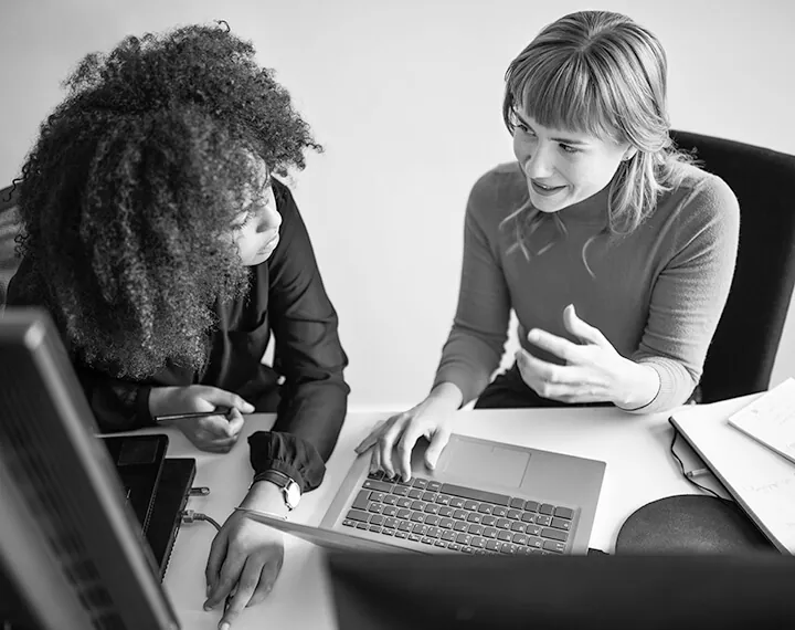 Two women engaged in discussion while seated at a desk, working together on a laptop.